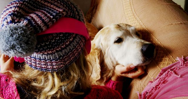 Child wearing a winter hat sits on the couch, enjoying a cozy moment with their golden retriever dog. Great for themes of companionship, warmth, home comfort, and pet love. Ideal for lifestyle blogs, pet-related content, and family-focused advertising.