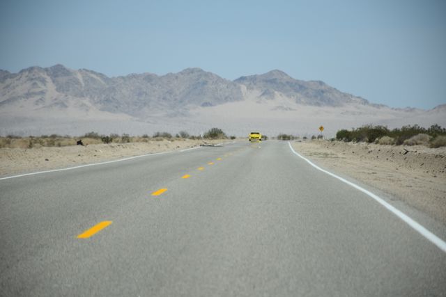 Lone yellow car driving down a stretch of deserted highway, with arid desert terrain and rugged mountains in the distance. This image captures the feeling of adventure and isolation, perfect for use in advertising travel, automotive products, or promoting road trips and scenic drives. Great for articles and marketing materials focusing on exploration and off-the-beaten-path travel.