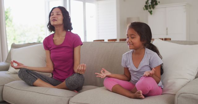Mother and daughter sitting on a couch in living room meditating and finding inner peace together. Perfect for themes on family bonding, mindfulness, wellness practices, mental health awareness, and peaceful home environments.