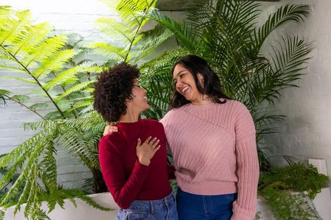 Smiling diverse couple embracing in bright conservatory with lush ferns and casual knitwear