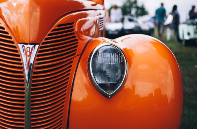 Close-up capturing the headlight and grille of a vintage orange car during an outdoor auto show. Suitable for websites and advertisements focusing on classic car enthusiasts, auto shows, vintage car exhibits, or automotive parts. Also ideal for illustrating nostalgic themes and retro designs.
