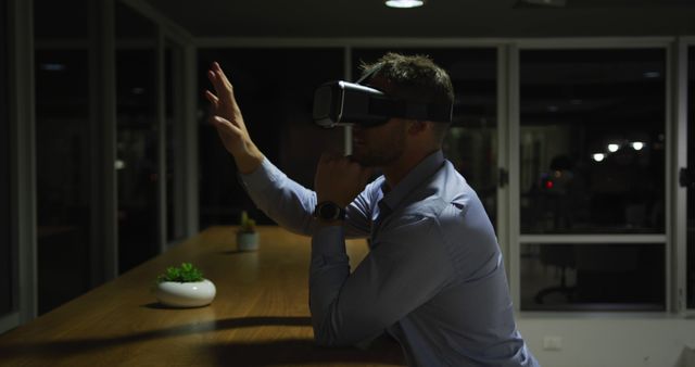 A man using a virtual reality headset indoors in a modern office. He is reaching out with his hand, engaging with the virtual world around him. This image can be used for articles and projects related to technology, business innovation, office environments, virtual reality, and modern tech advancements.