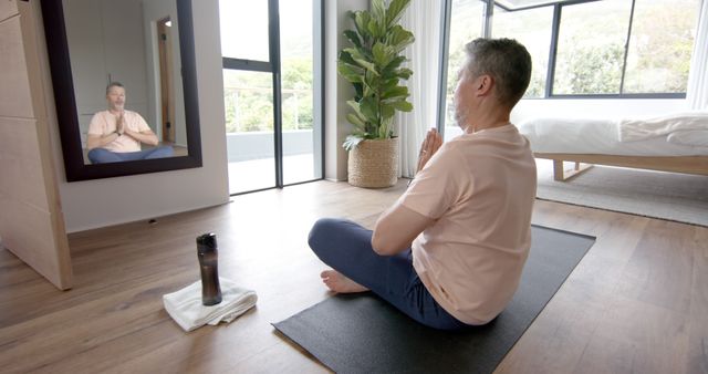 A middle-aged man is meditating in a modern home, sitting cross-legged on a yoga mat in front of a mirror. This serene image can be used for promoting mindfulness, wellness, and healthy lifestyles. Suitable for yoga studio ads, meditation apps, and wellness blogs.