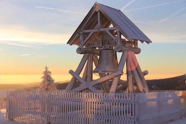 Frosted wooden bell tower in winter with a surrounding fence and snow-covered landscape during sunrise. This scene captures the tranquility and beauty of a cold morning in the mountains. Useful for promoting winter tourism, holiday themes, or architecture in nature visuals.