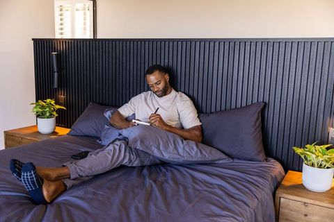 African american man writing in notebook on bed in minimalist cozy bedroom