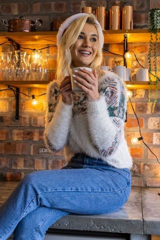 Smiling woman in festive sweater sipping coffee in rustic kitchen nook with string lights