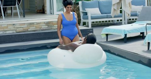 Indian woman in blue swimsuit smiling poolside while partner relaxing on inflatable float