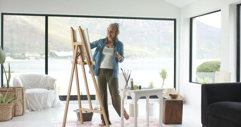 Mature woman painting at easel in sunlit lakeside living-room studio with scenic view