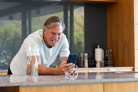 Mature man leaning on marble kitchen island smiling while checking smartphone