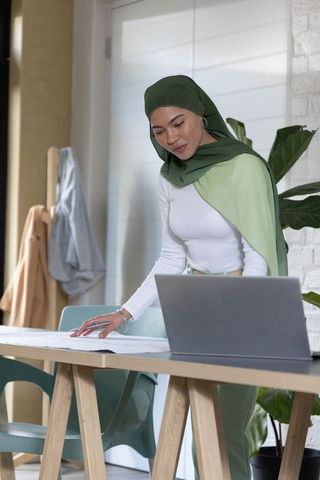 Woman wearing green hijab reviewing architectural plans at modern home studio workspace