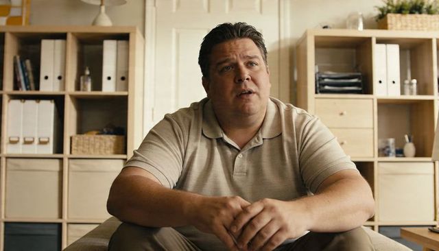 Middle-aged man sitting on couch wearing beige polo in cozy home office with shelving