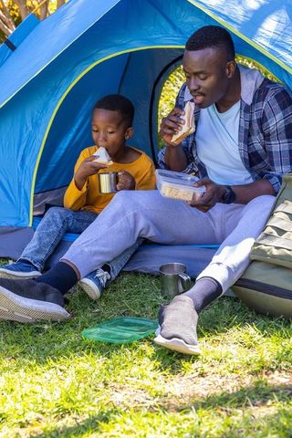 African american father and son enjoying sandwiches in blue tent while camping together