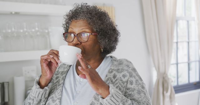 Senior African American woman with short curly hair and glasses drinks coffee in a bright kitchen setting. Suitable for themes around senior living, relaxation, home leisure activities, and morning routines.