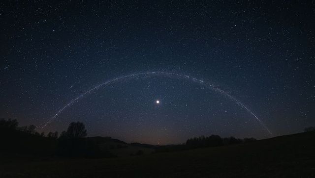 Semicircular star trail arch framing bright moon over rural field, milky way nightscape