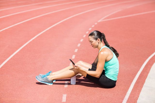 Female athlete sitting on running track using mobile phone. She is wearing athletic wear and appears to be resting after a workout. This image can be used for promoting fitness apps, sports gear, healthy lifestyle, or technology in sports.