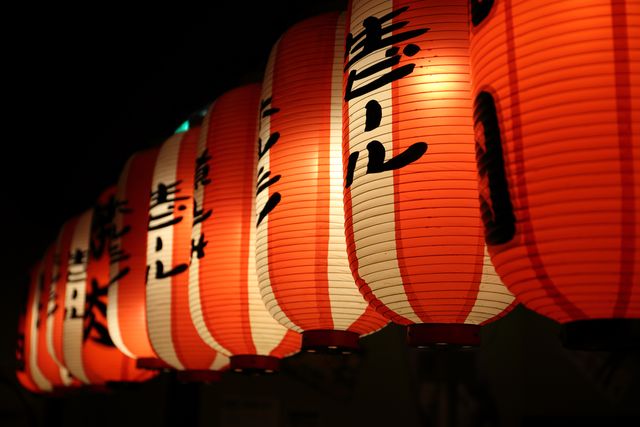 Traditional Japanese red paper lanterns with Kanji characters hanging in a row, well-lit and glowing at night. Useful for promoting cultural events, Japanese restaurants, travel promotions, and festivals. Creates a festive and cultural atmosphere reflecting the rich traditions of Japan.