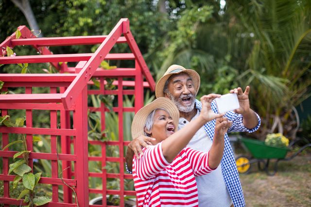 Elderly couple enjoying a moment together in a garden while taking a selfie. This image can be used in advertisements, articles, or blog posts focusing on active senior lifestyles, gardening, outdoor activities, and family memories. It evokes a sense of happiness, connection, and well-being.