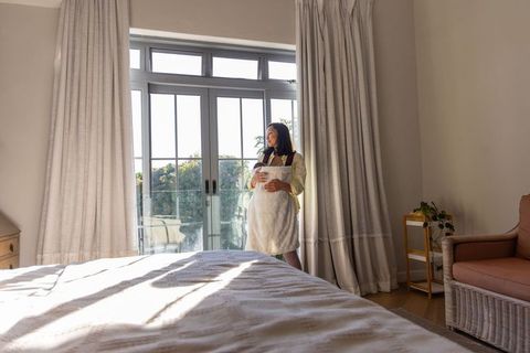 Pregnant woman standing by sunlit patio doors cradling belly in minimalist bedroom