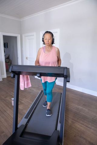 Senior asian woman walking on treadmill at home wearing headphones and activewear