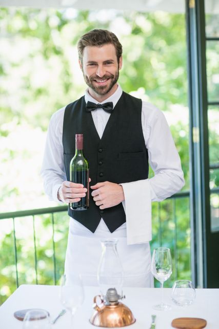 Smiling waiter holding a bottle of wine in a restaurant, dressed in a formal uniform with a bow tie and vest. Ideal for use in hospitality industry promotions, restaurant advertisements, customer service training materials, and dining experience visuals.
