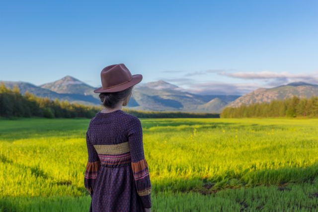 A woman stands in a large green field wearing a wide-brimmed hat, looking towards distant mountains under a clear blue sky. Ideal for advertisements promoting outdoor tourism, nature retreats, or adventure activities. Suitable for inspirational blog posts, travel brochures, and environmental awareness campaigns.