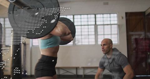 Female athlete lifting heavy barbell with coach spotting in industrial sunlit gym