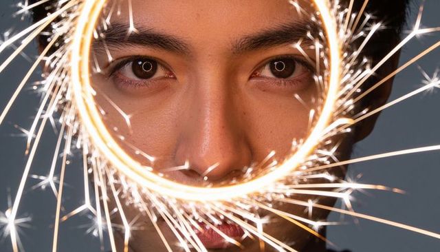 Asian man looking through circular sparkler, eyes reflecting ring light, dramatic closeup