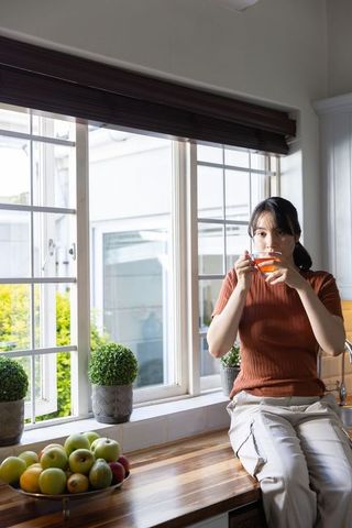 Asian woman sipping tea on kitchen counter by window with bowl of apples cozy lifestyle