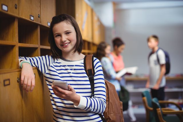 Schoolgirl standing in locker room, smiling while holding mobile phone. She is wearing a striped shirt and a backpack. Other students are seen in the background, engaging in conversation and reading. Ideal for use in educational materials, school promotions, technology in education, and youth lifestyle content.