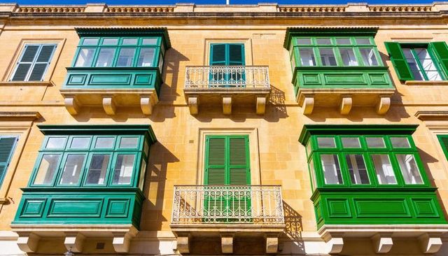 Sunlit mediterranean limestone facade featuring green enclosed balconies and iron railings