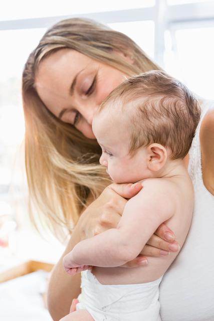 Woman holding her baby boy in her arms in a sunlit room. The image captures a moment of bonding and affection. Ideal for use in parenting blogs, family magazines, childcare advertisements, and product packaging for baby-related items.