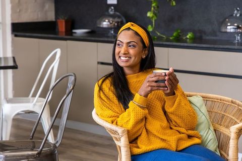 Smiling indian woman enjoying coffee in cozy mustard sweater and headband in cafe