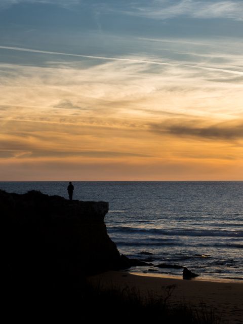 Person standing on cliff edge overlooking ocean horizon during sunset. Perfect for use in travel brochures, inspirational posters, websites related to nature or outdoor activities, and promotional material for coastal destinations.