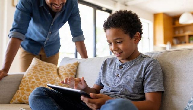 Smiling boy using tablet on sofa with caring parent nearby in bright cozy living room