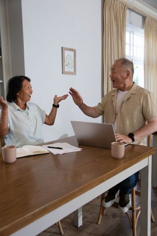 Senior chinese couple celebrating success at home desk high-fiving over laptop with coffee mugs