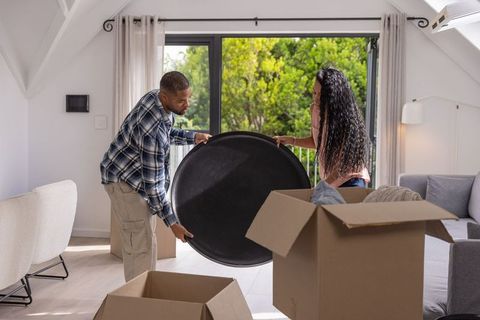 Young couple unpacking moving boxes carrying round tray in bright modern living room