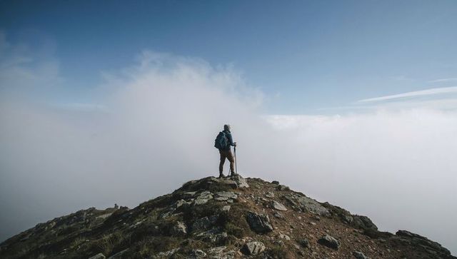 Standing hiker facing sea of clouds on rocky ridge carrying backpack and trekking pole