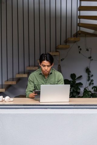 Asian man working from home on laptop and tablet in minimalist workspace with staircase plants