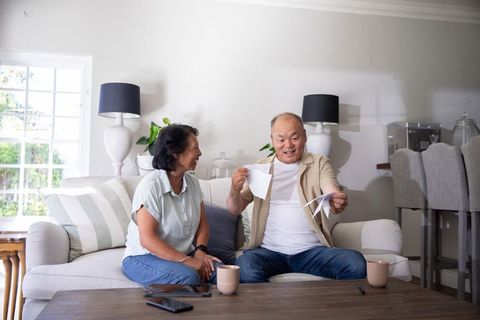 Senior asian couple tearing papers and smiling while sitting on cozy living room sofa