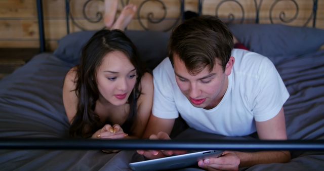 Couple is lying in bed together, sharing a digital tablet. Scene is cozy and casual, perfect for depicting modern relationships, technology use, and relaxation at home. Ideal for articles, blog posts, and advertisements focusing on lifestyle, home entertainment, and couple activities.