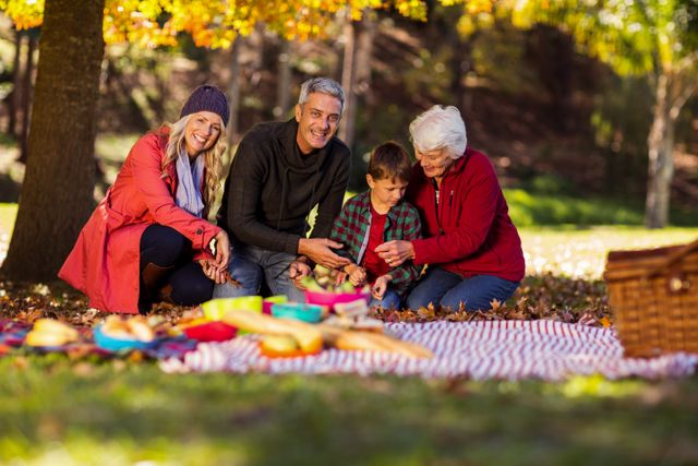 Multi-generation family enjoying a picnic in a park during autumn. The scene shows a senior, parents, and a child sitting on a blanket surrounded by colorful leaves. Ideal for use in advertisements, family-oriented content, and seasonal promotions highlighting family bonding, outdoor activities, and the beauty of autumn.