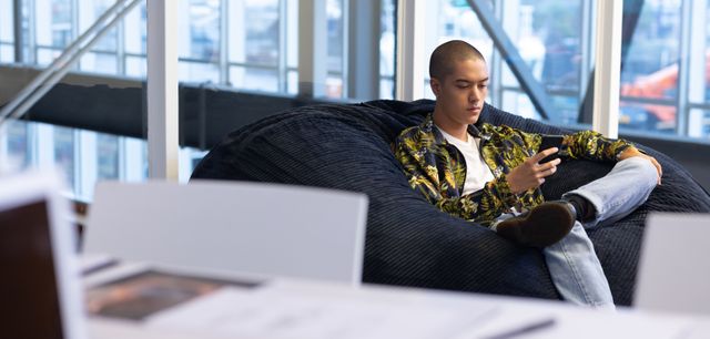 Young businessman sitting on a bean bag chair in a modern office, using a mobile phone. Ideal for illustrating modern work environments, technology use in business, relaxed office culture, and contemporary professional settings.