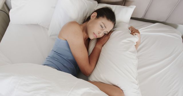 This image captures a woman lying in bed and cradling a pillow while wearing a worried expression, suggesting she may be dealing with insomnia or anxiety. Useful for articles and blogs on mental health, stress, sleep disorders, and nighttime routines. Also suitable for medical content, lifestyle magazines, and wellness promotions.