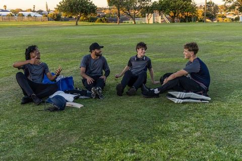 Young cricket teammates relaxing on grass field at golden hour sharing laughs and gear