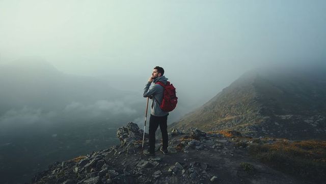 Solitary hiker standing on foggy mountain ridge with red backpack and trekking pole