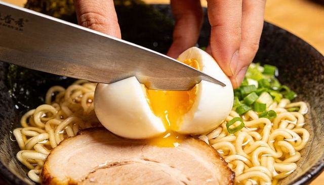 Slicing soft-boiled egg spilling golden yolk over steaming ramen with chashu and scallions