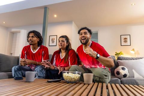 Friends cheering during soccer match at home on couch with popcorn and soccer ball