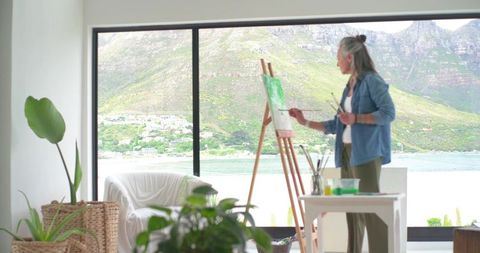 Mature woman painting coastal landscape at sunlit studio window, easel and brushes