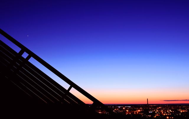 This image depicts a stunning sunset over an urban landscape with the silhouette of a railing on the left side. The sky transitions from deep blue to warm orange hues, highlighting the city lights below. Ideal for use in urban-themed projects, promotional materials for city tourism, or backgrounds for inspirational quotations.