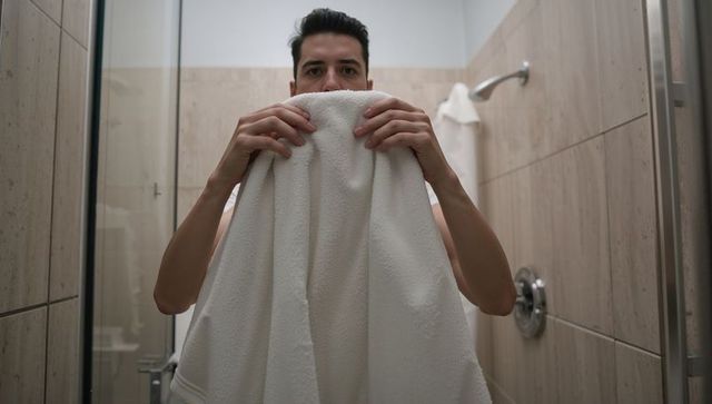 Young man holding large white towel covering lower face in modern beige shower stall